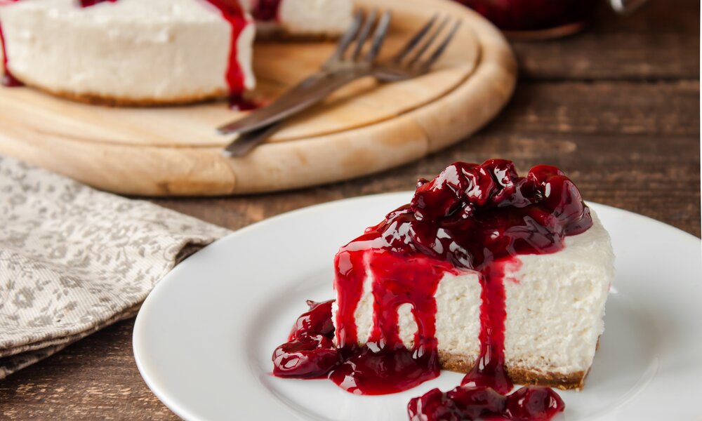 Stück Käsekuchen mit Kirschkompott auf einem Teller, dahinter der gesamte Kuchen. | © Adobe Stock / yuliiaholovchenko