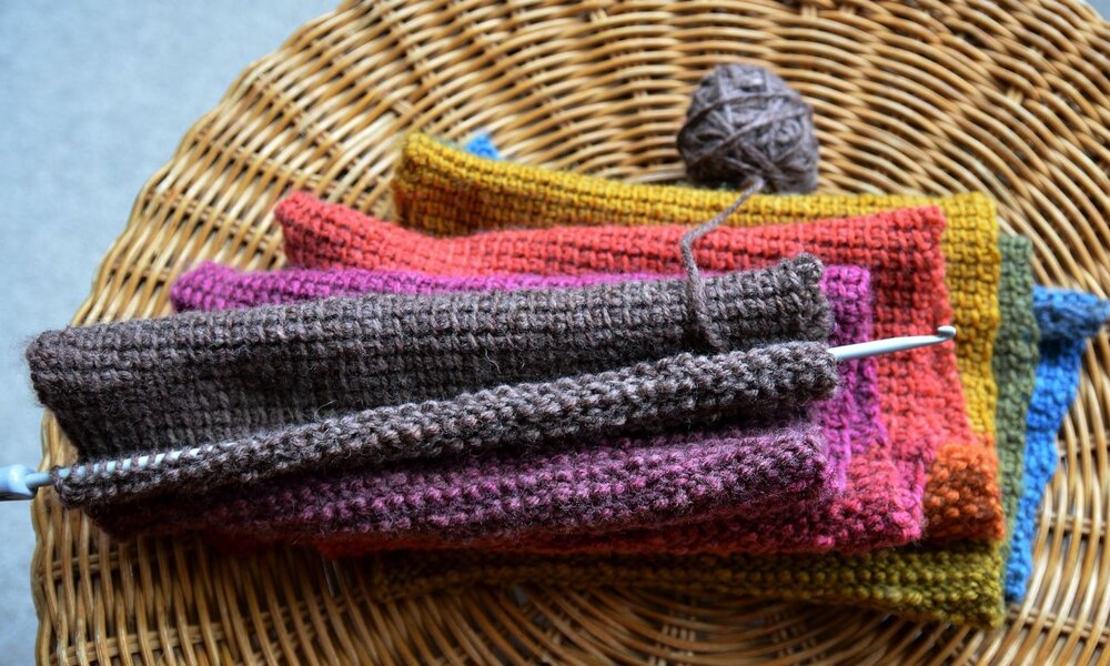 Mehrere gestrickte Stoffstücke in kräftigen Farben liegen ordentlich auf einem runden Korbtisch, darüber eine angefangene braune Strickarbeit. | © kate / stock.adobe.com