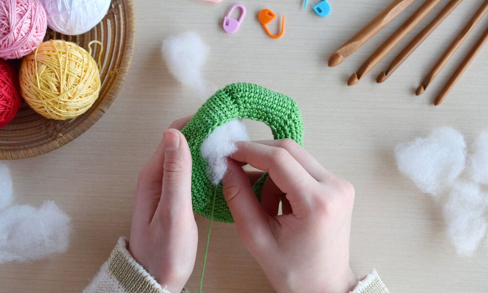 Nahaufnahme von Händen, die ein gehäkeltes, grünes Werkstück mit Watte füllen, umgeben von buntem Garn und Häkelnadeln. | © Oksana_S / stock.adobe.com