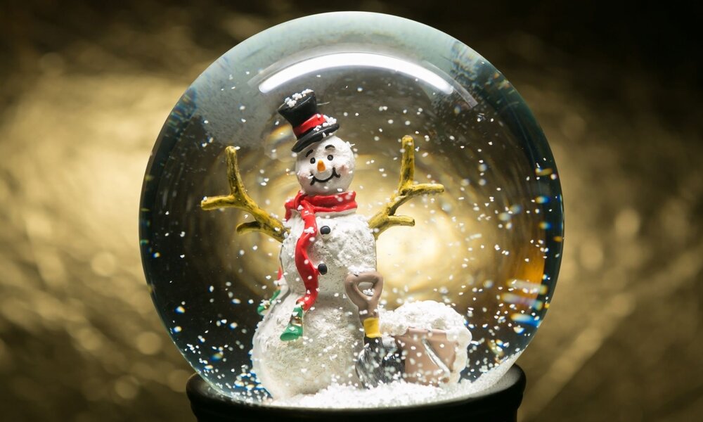 Schneemann mit rotem Schal und schwarzem Zylinder in einer Schneekugel vor goldenem Hintergrund, umgeben von wirbelnden Schneeflocken. | © Balazs/stock.adobe.com