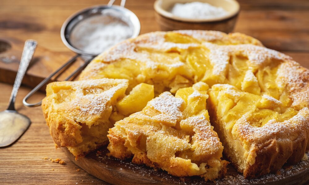 Angeschnittener Apfelkuchen in Kuchenform, daneben frische Äpfel. | © Adobe Stock / O.B.