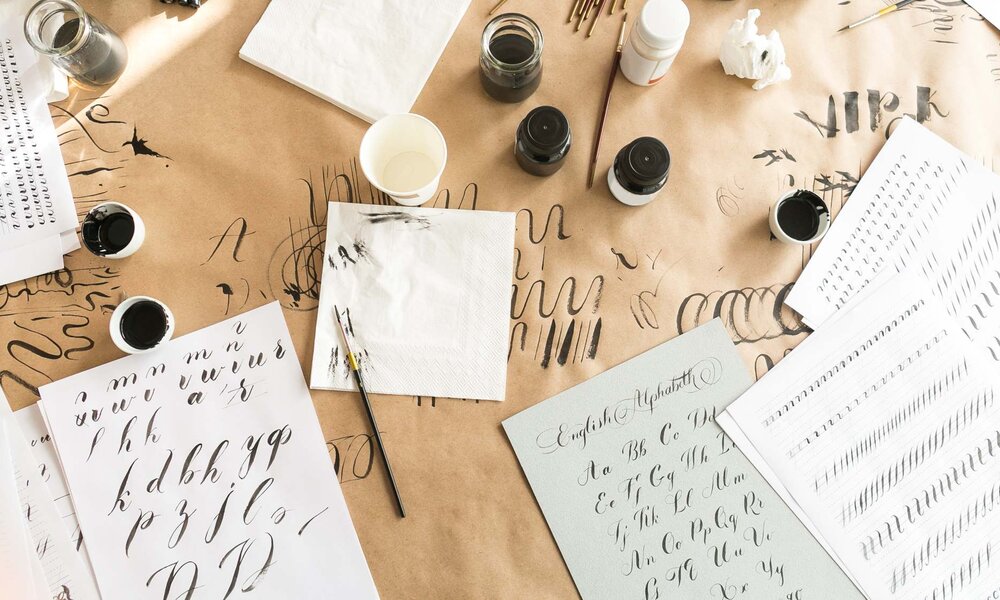 Kalligrafie-Arbeitsplatz mit Papierbögen, auf denen elegante Buchstaben geübt werden, sowie verstreuten Tintenbehältern und Werkzeugen. | © melnikofd/stock.adobe.com 