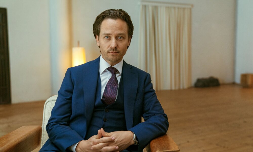 Tom Schilling als Björn Diemel sitzt ernst blickend auf einem Holzstuhl in einem minimalistischen Raum. | © Netflix / Achtsam Morden