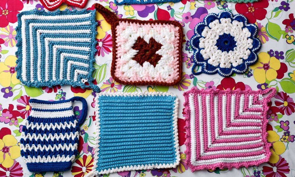 Bunte handgehäkelte Topflappen in verschiedenen Mustern und Farben liegen auf einer Tischdecke mit lebhaftem Blumenmuster. | © Mamo/stock.adobe.com