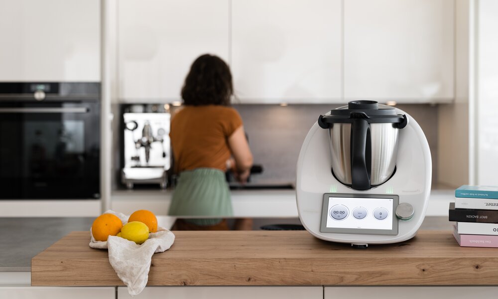 Thermomix auf Holzarbeitsfläche mit Zitrusfrüchten, im Hintergrund kocht eine Frau mit braunen Locken in einer hellen Küche. | © Vorwerk