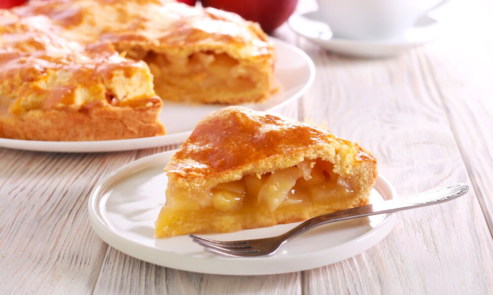 Stück Apfelkuchen vor einem ganzen Kuchen auf einem Tisch. | © Adobe Stock / manyakotic