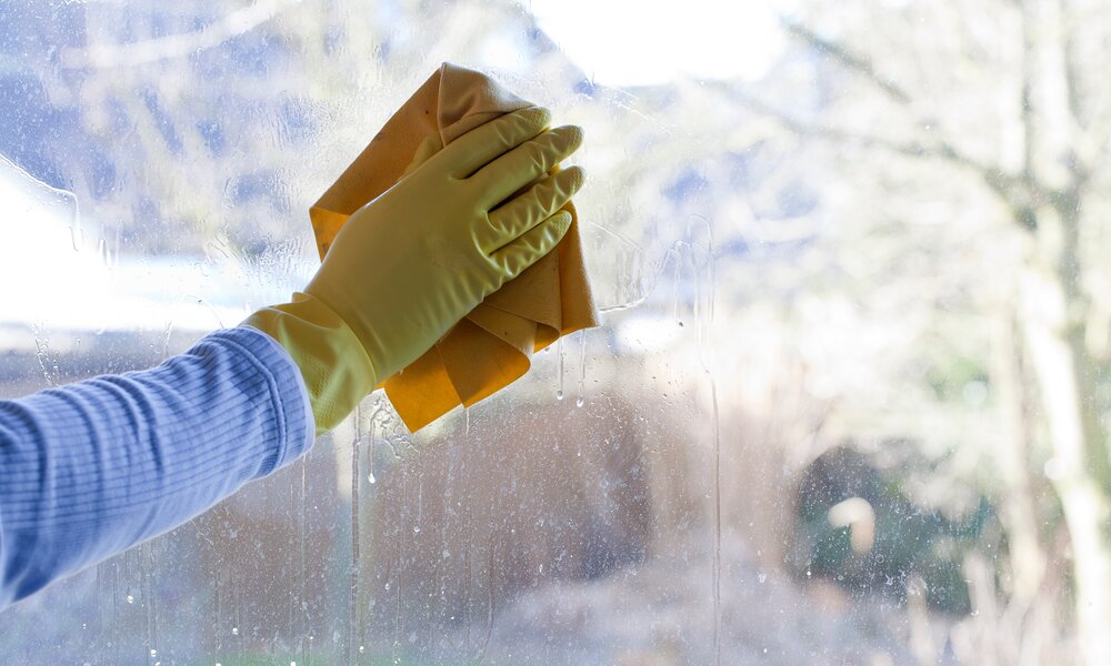Dreckiges und fleckiges Fenster, im Hintergrund Garten, links blauer Arm mit gelben Handschuh über Hand, putzt das Fenster mit gelben Tuch | © Adobe Stock/D. Ott