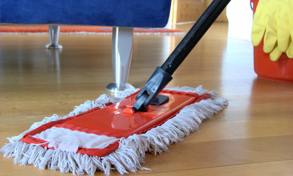 Holzboden, im Hintergrund Füße von blauem Sofa, auf Boden Bodenwischer mit weißem Bezug, im Hintergrund roter Eimer mit Handschuhen drüber | © Adobe Stock/Marina Lohrbach