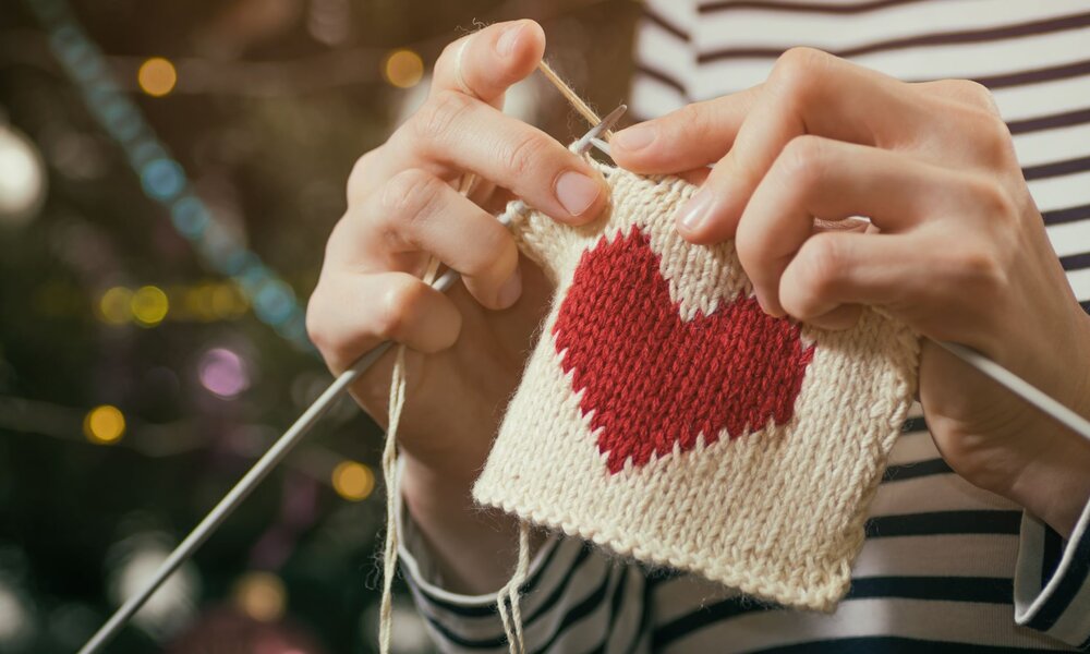Nahaufnahme von strickenden Händen, die ein rotes Herz auf weißem Strickstück fertigen, vor festlichem Hintergrund. | © svetazi/stock.adobe.com