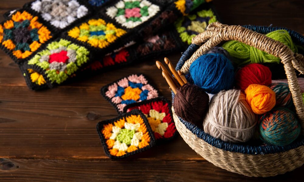 Ein Korb mit bunten Wollknäueln und Häkelnadeln steht auf einem Holztisch, daneben leuchtende, gehäkelte Granny-Squares. | © Tsurukame Design/stock.adobe.com