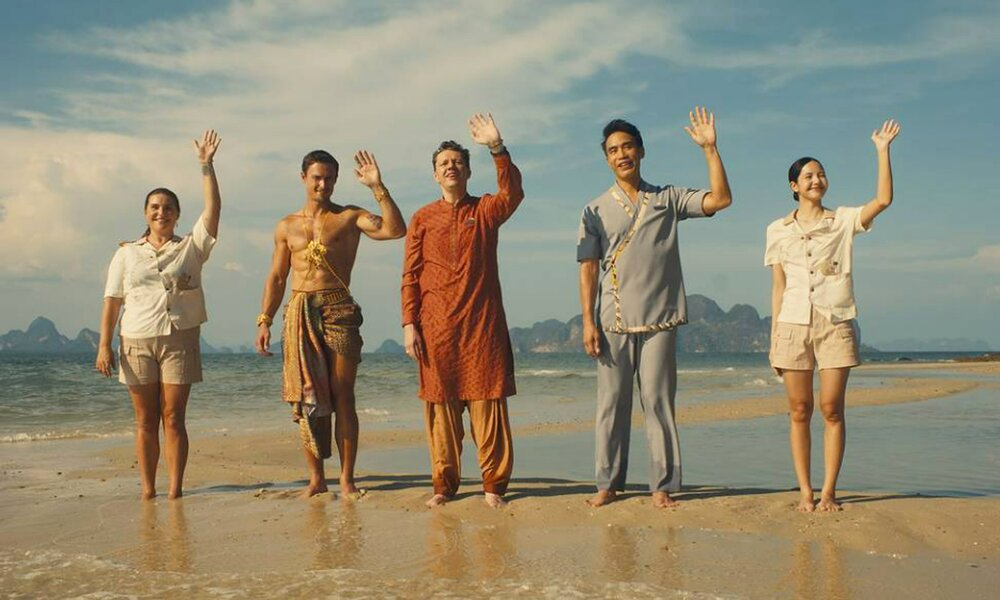 Die Crew des Resort aus  der dritten Staffel der HBO-Serie "White Lotus" steht winkend am Strand im seichten Wasser unter blauem Himmel | © HBO/Sky