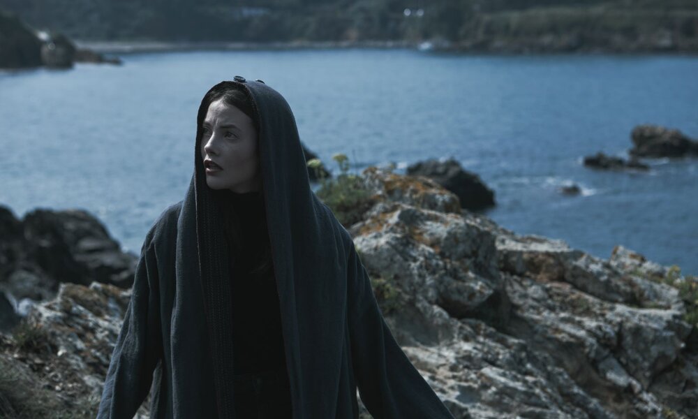 MIREIA ORIOL as ALMA in episode 07 of ALMA: Junge Frau mit heller Haut und dunklen Haaren im langen, grauen Umhang blickt besorgt auf felsiger Küste mit blauem Meer. | © ALMA LARRAÑAGA/NETFLIX