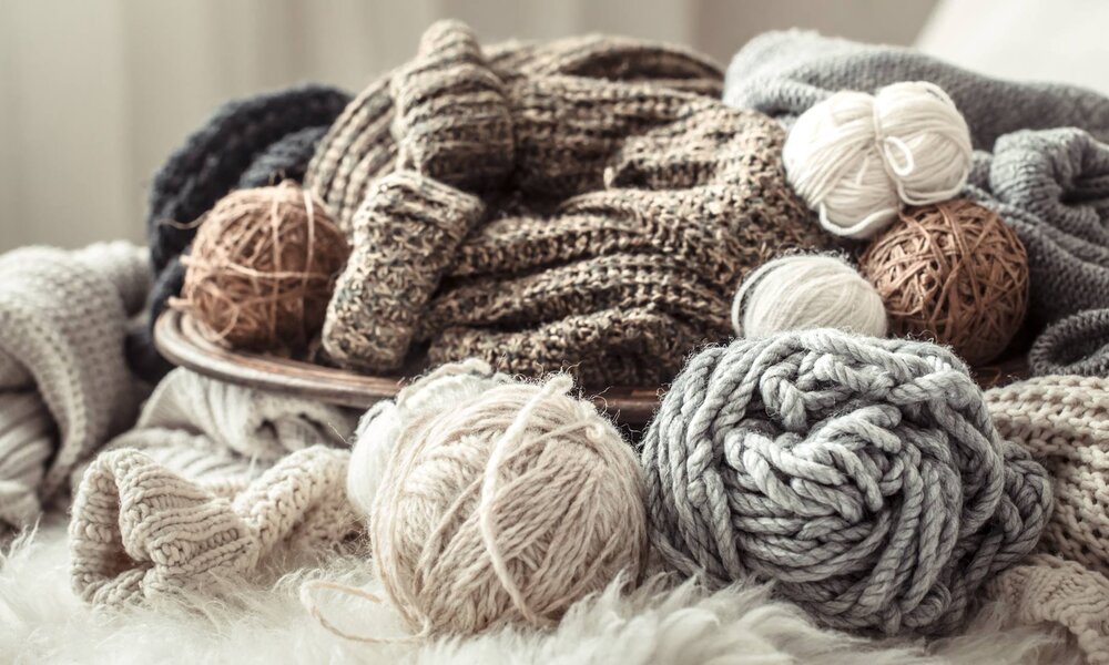 Verschiedene Wollknäuel und gestrickte Kleidungsstücke in sanften Naturfarben sind auf flauschigem Untergrund arrangiert. | © puhimec/stock.adobe.com