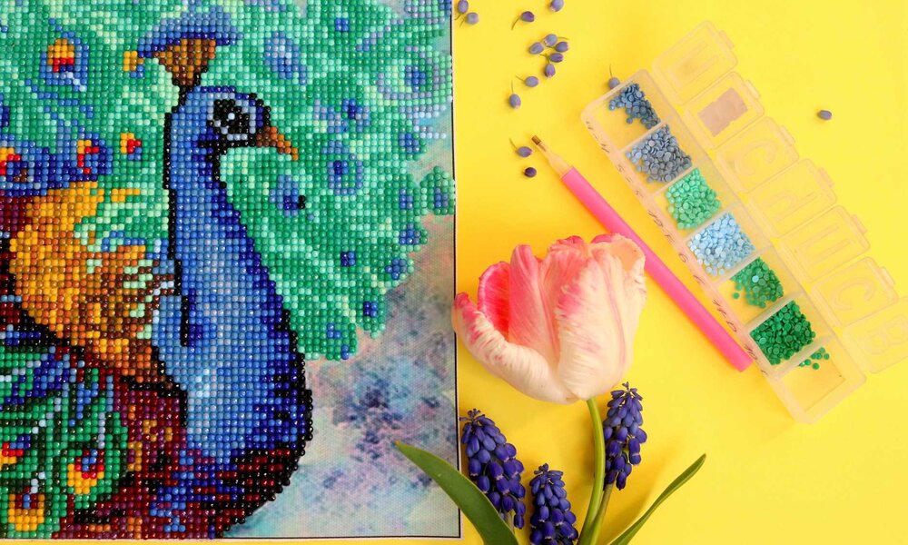 Detailreiches Diamond-Painting eines blauen Pfaus, umgeben von grünen und blauen Strasssteinen, mit Tulpe und Werkzeug auf gelbem Hintergrund. | © Olga/stock.adobe.com