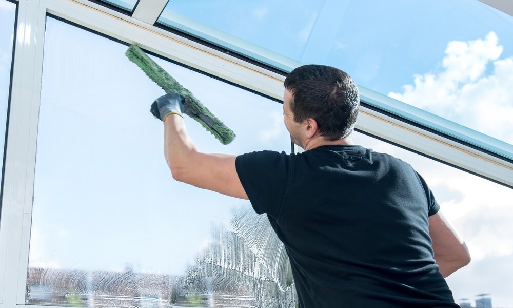 Mann mit schwarzem Shirt steht an großer Fensterscheibe mit Blick in den Garten, er putzt sie mit einem Wasserabzieher, Wetter draußen ist gut und sonnig | © Adobe Stock/pictworks