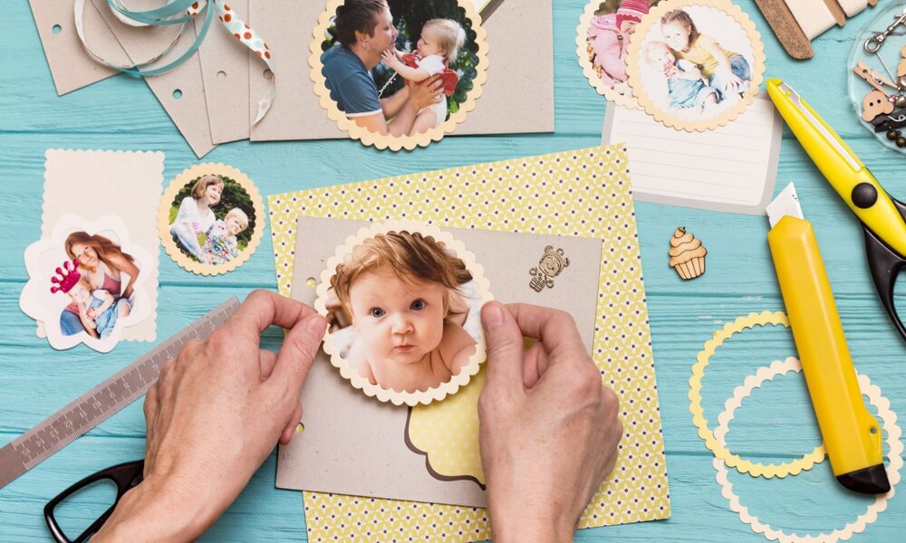 Hände gestalten ein Scrapbook auf blauem Holztisch, dekoriert mit ausgeschnittenen Babyfotos, Bastelpapier, Schere und Cutter. | © Sergii Mostovyi /stock.adobe.com
