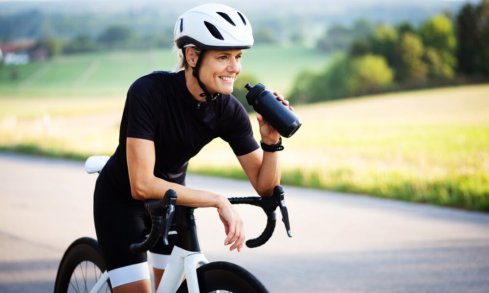 Frau auf Rennrad mit weißem Helm und schwarzen Outfit, in der Hand eine Trinkflasche aus der sie gerade trinken will, Hintergrund ist ländlich mit grüner Wiese | © Adobe Stock/Wellnhofer Designs