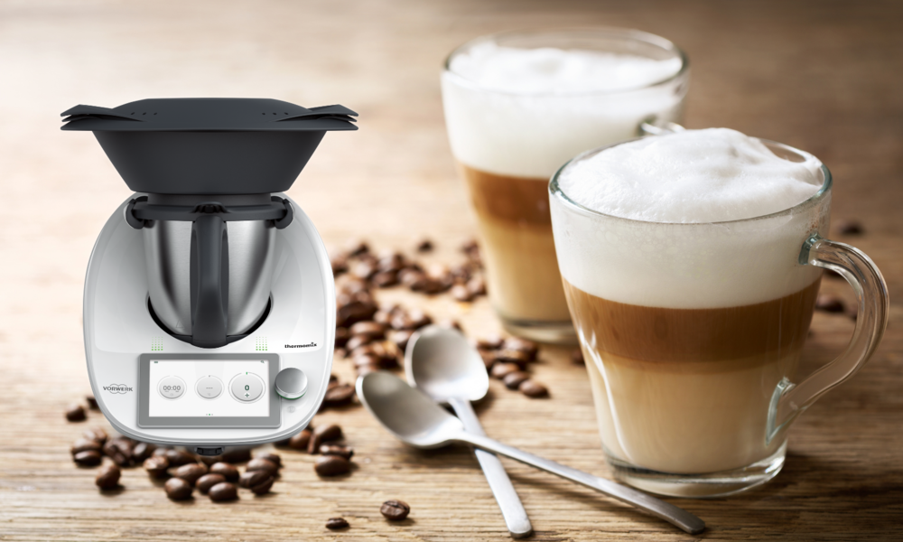 Weißer Thermomix vor einer rustikalen Holzplatte, flankiert von zwei Latte-Macchiato-Gläsern mit dicker Milchschaumkrone und Kaffeebohnen. | © Adobe Stock/ Nitr / Vorwerk