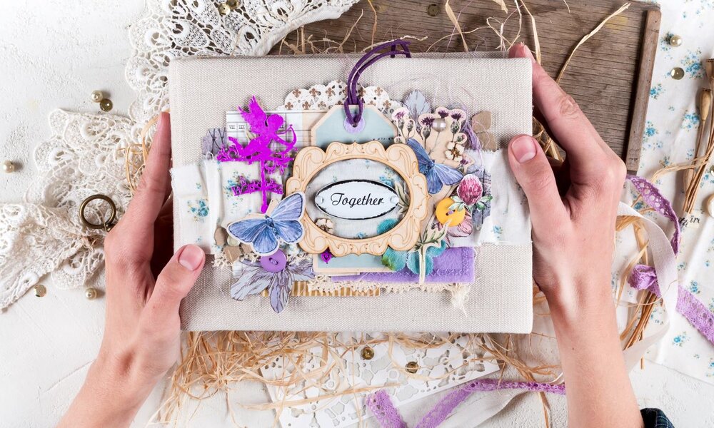 Zwei Hände halten ein kunstvoll verziertes Scrapbook mit floralen Details, Spitze und dem Schriftzug „Together“ in elegantem Rahmen. | © Svetlana Glazkova /stock.adobe.com