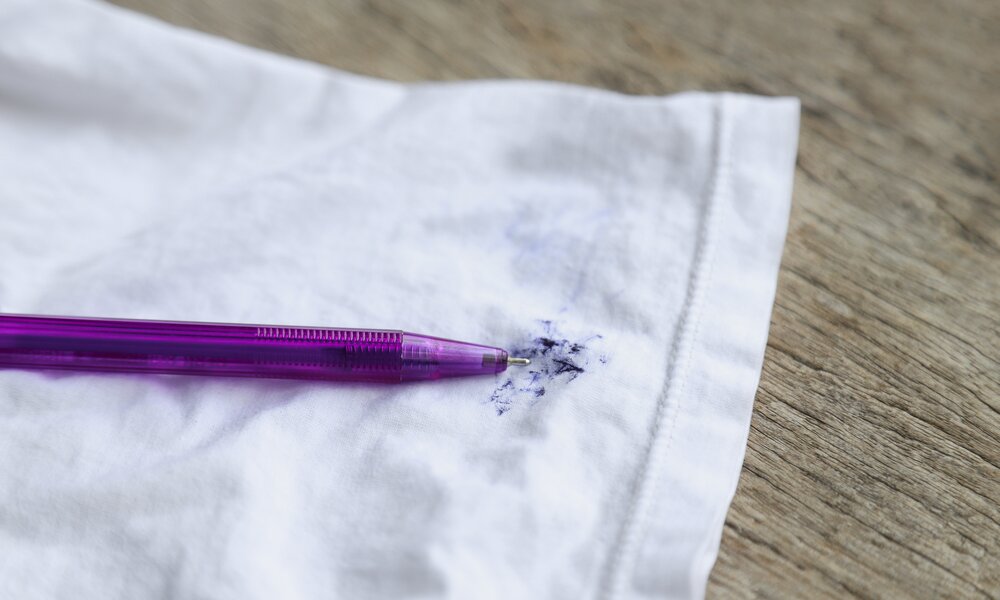 Ein lila Kugelschreiber liegt auf einem weißen Stoff, der mit blauer Tinte befleckt ist, auf einer Holzoberfläche. | © Adobe Stock/ Apiwat