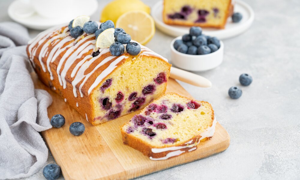 Angeschnittener Blaubeer-Kuchen auf einem Brett, daneben und dahinter frische Blaubeeren und Zitronen | © Getty Images / 	Irina Taskova