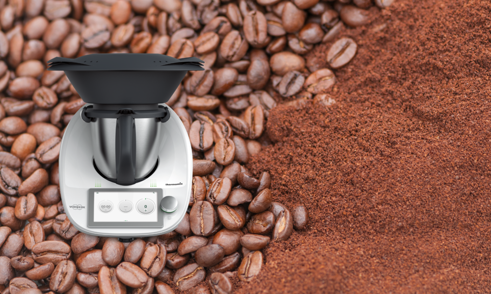 Nahaufnahme eines Thermomix vor braunen Kaffeebohnen und einer Fläche aus gemahlenem Kaffee. | © Adobe Stock/ mahey/ Vorwerk