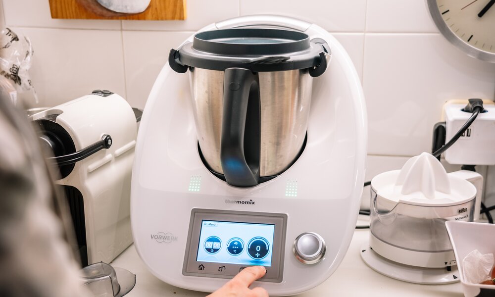 Weißer Thermomix TM6 mit glänzendem Edelstahlbehälter steht in heller Küche, daneben eine Zitruspresse und Kaffeemaschine. | © Adobe Stock/ Guillem de Balanzó