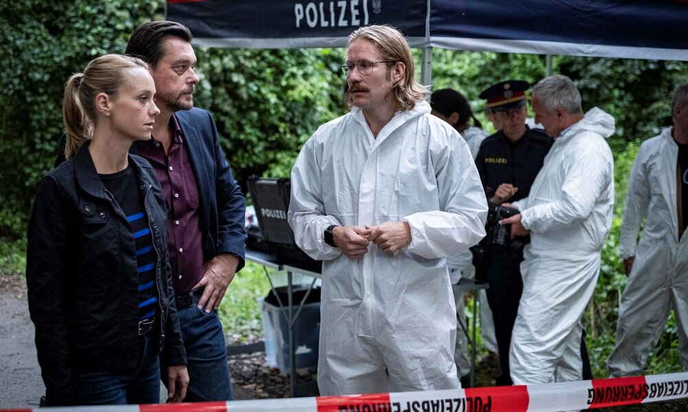 Eine blonde Polizistin und ein dunkelhaariger Mann in Zivil sprechen mit einem Forensiker in Schutzanzug an einem abgesperrten Tatort im Wald. | © ARD Degeto Film/Allegro Film/Toni Muhr