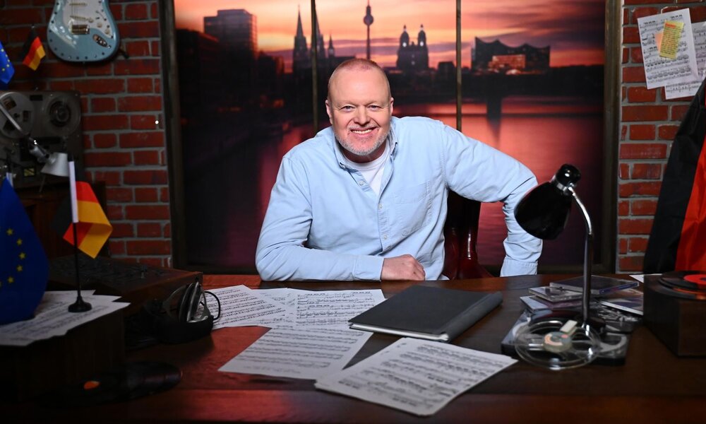 Stefan Raab in hellblauem Hemd sitzt entspannt an einem Schreibtisch mit Notenblättern, Tonband und Flaggen. | © RTL / Raab Entertainment / Julia Feldhagen