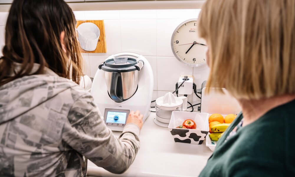 Eine junge Frau in grauer Kapuzenjacke bedient einen Thermomix in einer hellen Küche, während eine blonde Frau zusieht. | © Adobe Stock/ Guillem de Balanzó