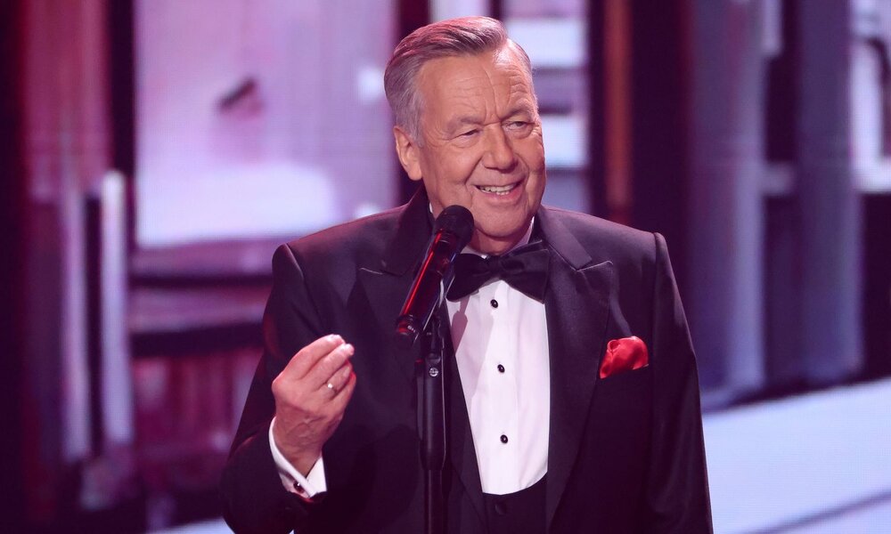 Ein älterer Mann im schwarzen Smoking mit rotem Einstecktuch singt lächelnd in ein Mikrofon auf einer beleuchteten Bühne. | © Getty Images / Gerald Matzka