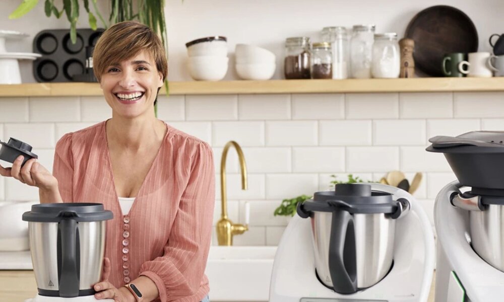 1. Eine lächelnde Frau mit kurzem braunem Haar steht in einer Küche hinter drei Thermomix-Modellen, umgeben von Pflanzen und Regalen.    2. Junge Frau in altrosa Bluse präsentiert drei moderne Thermomix-Geräte auf einer hellen Küchenarbeitsplatte.    3. In einer hellen Küche zeigt eine Frau mit kurzem Haar und Schmuck am Arm stolz drei verschiedene Thermomix-Modelle.   | © Vorwerk