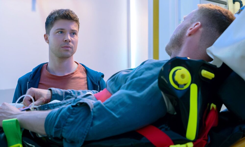 Ein bewusstloser Mann mit blondem Haar liegt fixiert auf einer Trage, während ein junger Mann mit ernster Miene danebensteht. | © MDR/Saxonia Media/Sebastian Kiss