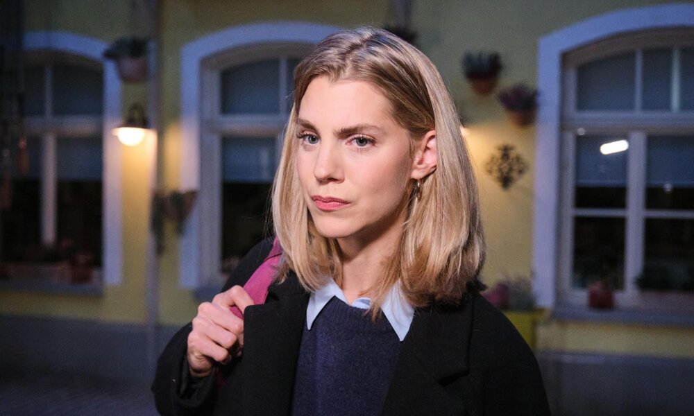Junge blonde Frau mit ernstem Blick trägt eine schwarze Jacke und einen rosa Rucksack vor einer beleuchteten Hausfassade am Abend. | © RTL / Stefan Behrens