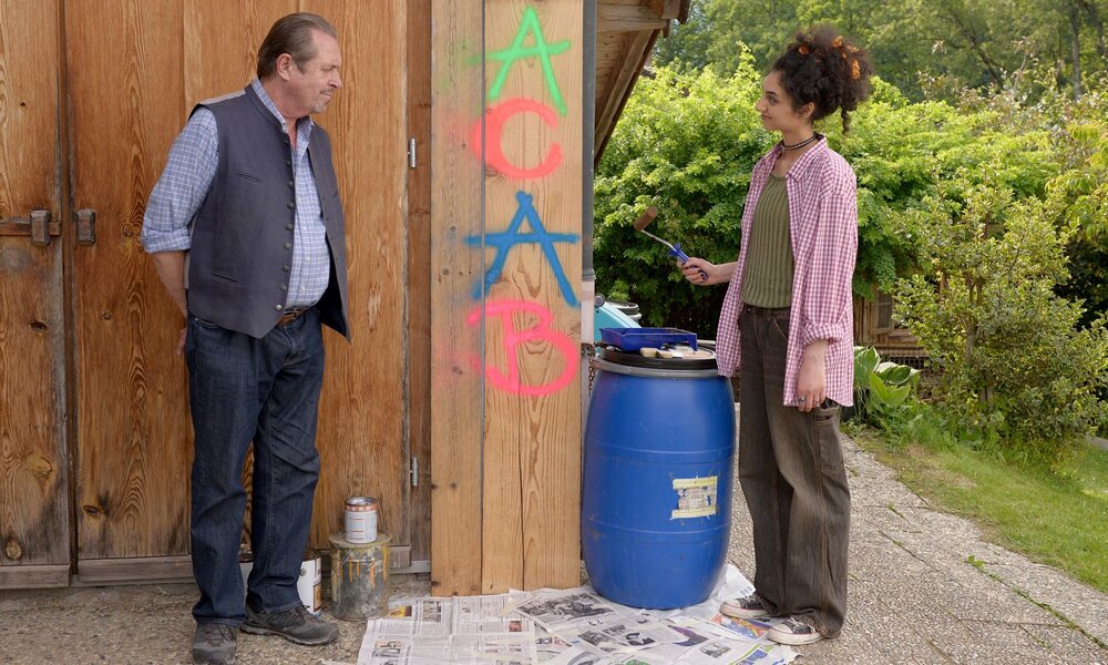 Ein älterer Mann in Trachtenweste blickt skeptisch auf eine junge Frau mit Locken, die eine Wand mit buntem Graffiti besprüht. | © ARD/Rupert Scheele