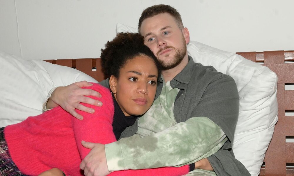 Eine junge Frau in pinkem Pullover liegt besorgt im Arm eines nachdenklichen Mannes mit Bart auf einem Bett mit weißen Kissen. | © RTL / Rolf Baumgartner