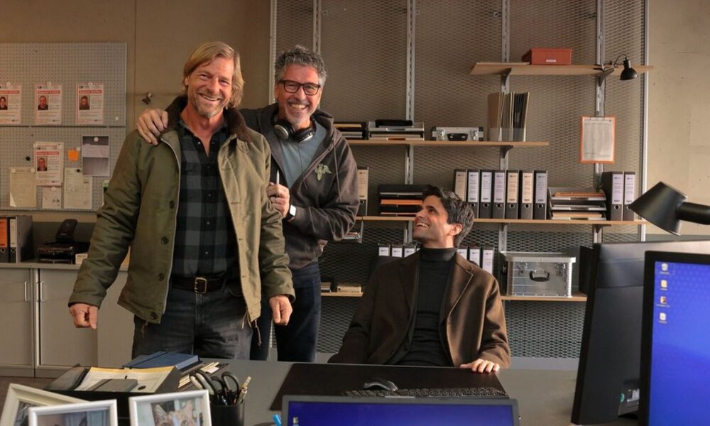 In einem modern eingerichteten Büro mit Regalen voller Aktenordner stehen zwei Männer lachend nebeneinander, während ein dritter im Sessel sitzt und zu ihnen aufblickt. | © SAT.1/Tom Trambow