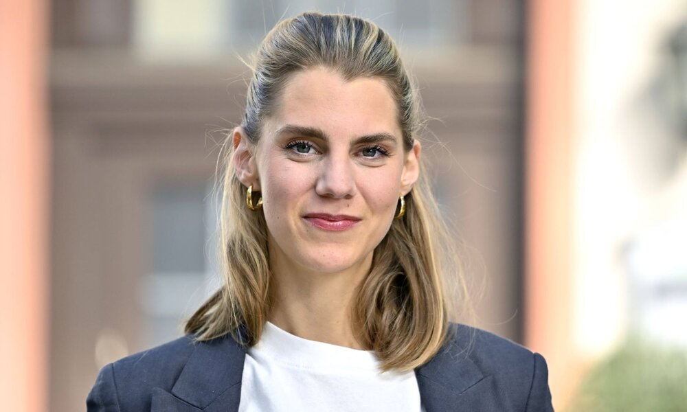 Eine junge Frau mit schulterlangem blondem Haar, goldenen Ohrringen und dunklem Blazer lächelt selbstbewusst vor unscharfem Stadt-Hintergrund. | © IMAGO Images / Future Image / D. Anoraganingrum