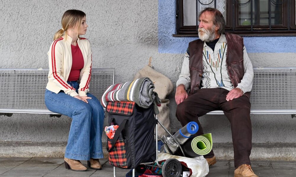 Eine junge Frau in Jeans und Sportjacke sitzt auf einer Bank und blickt nachdenklich zu einem älteren, bärtigen Obdachlosen mit Habseligkeiten. | © ZDF/Barbara Bauriedl