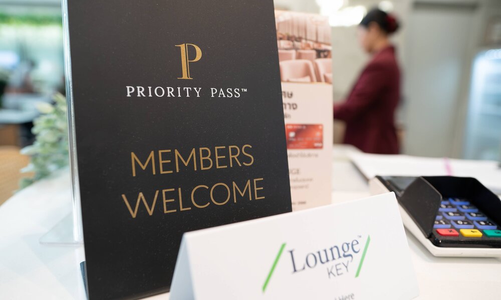 Ein schwarzes Schild mit goldener Aufschrift „Priority Pass – Members Welcome“ steht auf einem weißen Tresen in einer Flughafenlounge. | © IMAGO/ZUMA Press Wire