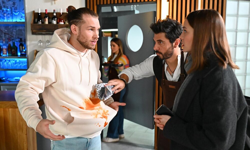 Ein junger Mann mit Dutt und cremefarbener Kapuzenjacke blickt verärgert auf seinen mit Tomatensauce bekleckerten Pullover, während ein Kellner mit dunklen Haaren hektisch versucht, den Fleck mit einem Tuch zu entfernen. | © RTL / Julia Feldhagen