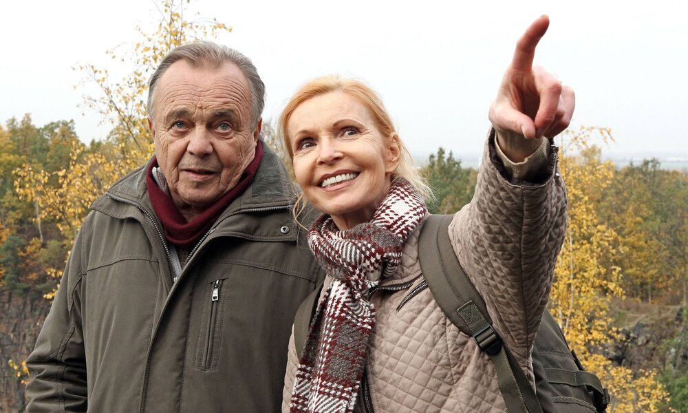Ein älteres Paar in warmen Jacken steht vor buntem Herbstlaub, die Frau strahlt und deutet auf etwas, während der Mann interessiert blickt. | © IMAGO Images / STAR-MEDIA