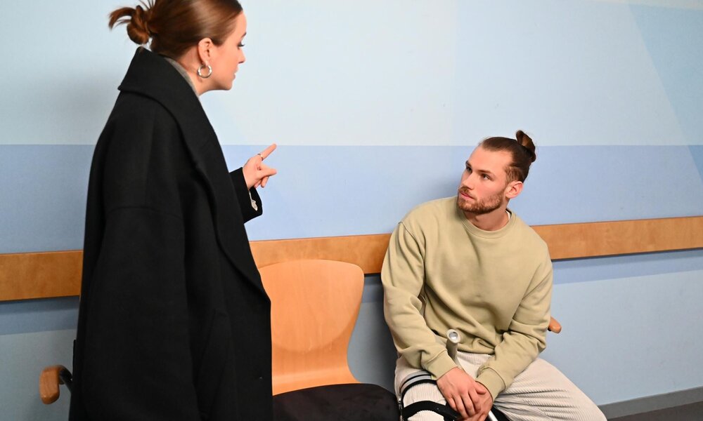 Eine junge Frau mit dunklem Mantel und Dutt spricht energisch mit einem sitzenden jungen Mann mit Zopf und Krücken im Krankenhausflur. | © RTL / Julia Feldhagen