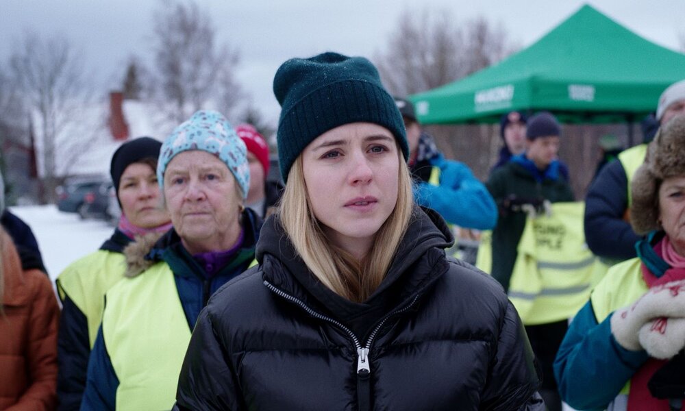 Bild der Netflix-Krimiserie "Die Åre-Morde": Hanna Ahlander blickt ernst in die Ferne, umgeben von Menschen in Winterkleidung. | © 2024 Netflix, Inc