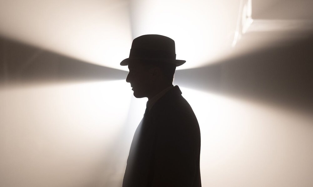 Ein Mann im Profil mit Hut im Retro-Stil steht vor hellem Licht, das Schatten und Strahlen auf die Wand projiziert. | © Frédéric Batier/X Filme Creative Pool/ARD Degeto Film/SWR/WDR/Radio Bremen/Beta Film 