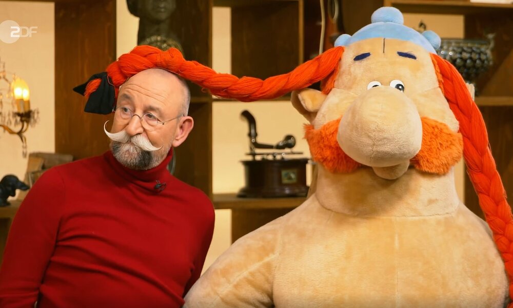 Ein Mann mit grauem Bart in rotem Pullover posiert mit Asterix-Figur, deren roter Zopf auf seinem Kopf liegt. | © ZDF