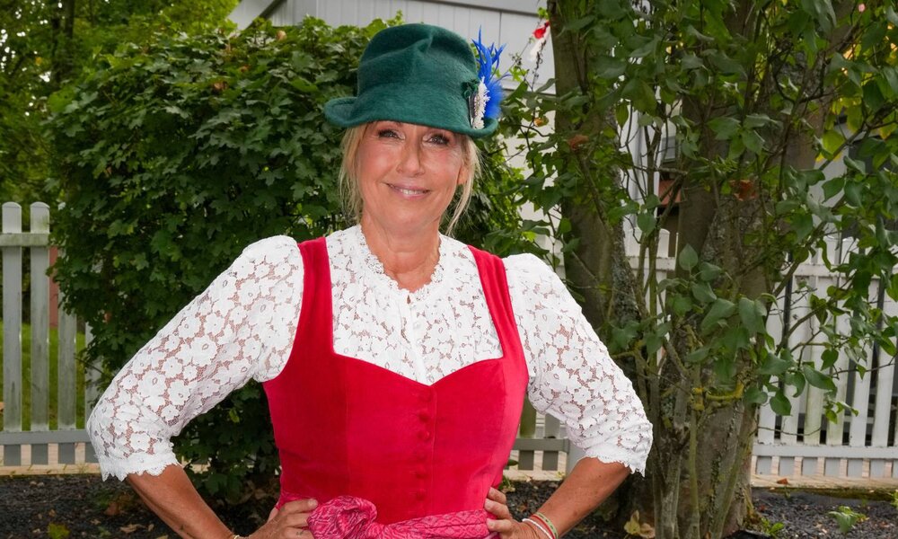 Eine blonde Frau in rotem Dirndl mit weißer Spitzenbluse und grünem Trachtenhut lächelt selbstbewusst vor einem Gartenzaun. | © IMAGO / BOBO