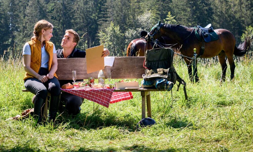 Ein Paar genießt ein Picknick mit Sekt auf einer Wiese, umgeben von Wäldern, während ihre gesattelten Pferde im Hintergrund grasen. | © ARD/WDR/Christof Arnold