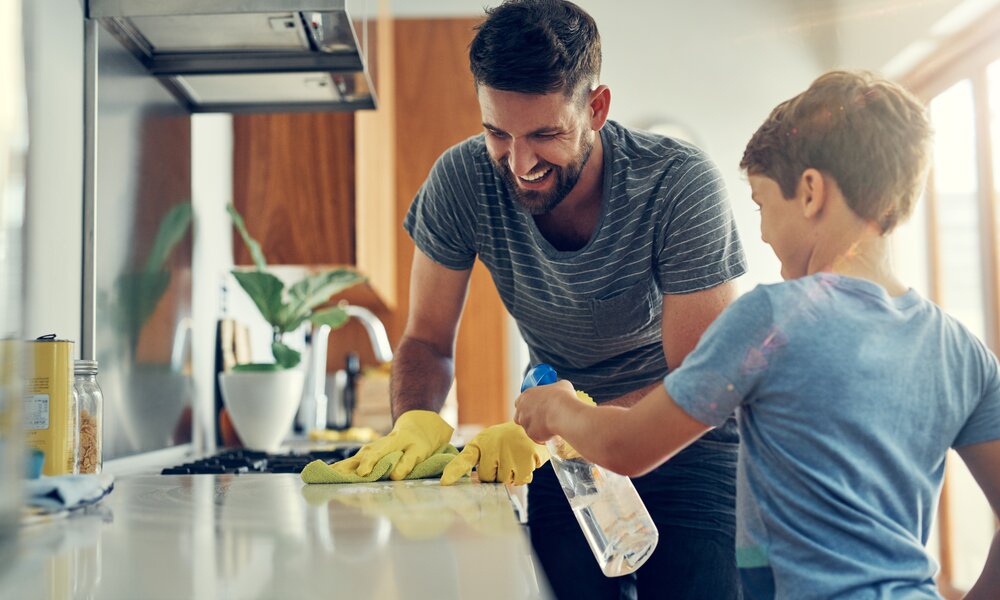Ein lachender Vater mit Bart und gestreiftem Shirt reinigt mit gelben Handschuhen die Küchenarbeitsplatte, während sein Sohn hilft. | © Adobe Stock/ Nikish Hiraman/peopleimages.com