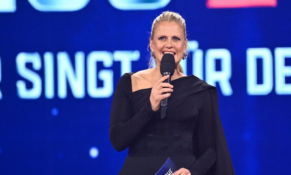 Eine blonde Frau in elegantem schwarzem Kleid moderiert mit Mikrofon lächelnd auf einer blau beleuchteten Bühne. | © RTL / Raab Entertainment / Willi Weber